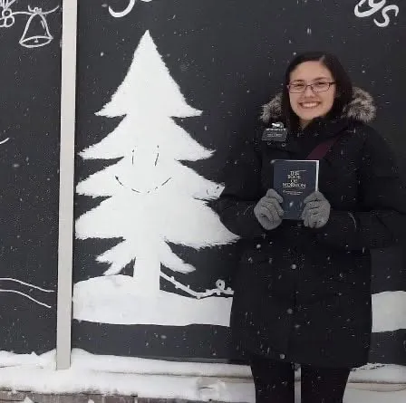 Rachel in a winter coat holding a Book of Mormon beside a painted white Christmas tree in the snow